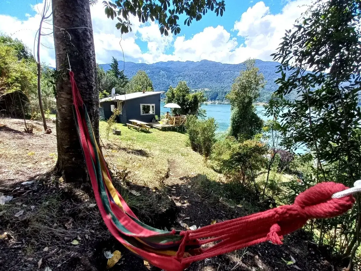 Hamaca roja entre los árboles con vista a una cabaña, el agua y las montañas